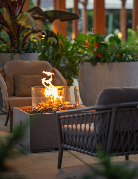 Outdoor patio with cushioned chairs around a glass-enclosed fire pit, surrounded by large potted plants and wooden beams.